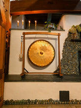 Load image into Gallery viewer, Golden gong on its wooden stand inside Wild Indigo Forest Farm's artisan-built cob and stone retreat space, illuminated by candles.