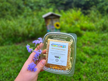 Load image into Gallery viewer, Wild Indigo Forest Farm raw comb honey held beside purple flowers with the real grassy apiary background
